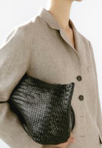 Pochette noire tissée tenue contre une chemise beige clair boutonnée. Le sac présente une apparence douce et texturée, mettant en valeur son design unique.