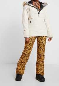 Weiße wasserdichte Jacke mit schwarzem Innenfutter und Kapuze mit Fellbesatz, kombiniert mit orangen Schneehosen im Leopardenmuster und schwarzen Stiefeln.