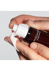 Brun glasflaska med vit pump som dispenserar en krämig vit lotion på ett fingertopp; slät textur, minimalistisk design.