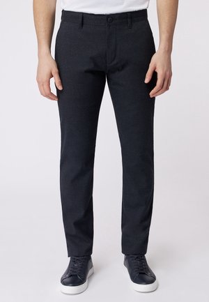 Mand iført slim-fit mørkegrå bukser og sorte sneakers med hvide såler, stående foran en ensartet lys baggrund.