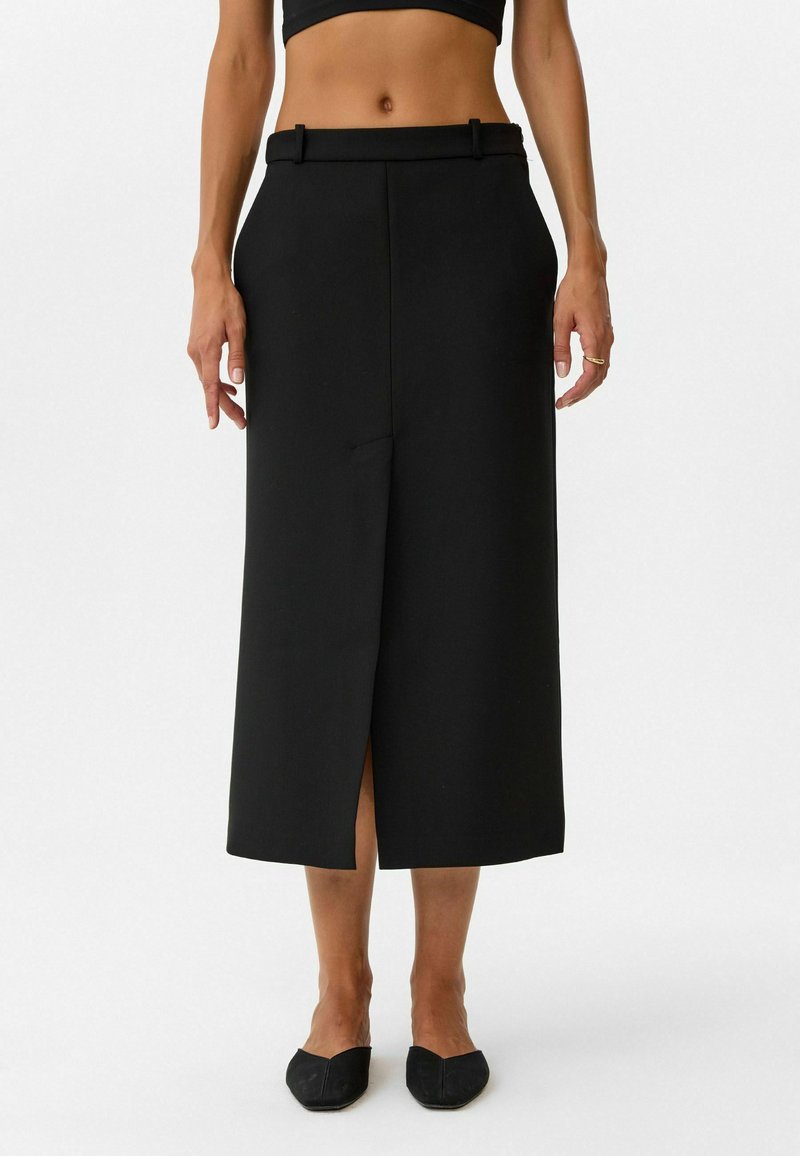 Schwarze Midikleid mit geradem Schnitt, mit Schlitz vorne, Gürtelschlaufen und zwei Side-Pockets. Hergestellt aus glattem Stoff, kombiniert mit schwarzen flachen Schuhen.