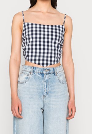 Kobieta ubrana w czarno-biały kraciasty, krótki top na cienkich ramiączkach oraz jasnoniebieskie, dopasowane jeansy z wysokim stanem.
