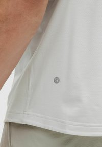 Camiseta deportiva de color gris claro con tejido texturizado y transpirable con pequeñas perforaciones, acentuada por un logo plateado en el lado inferior.