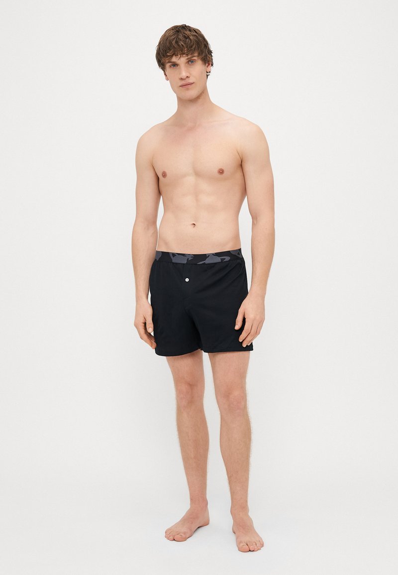 Schwarze Badeshorts mit einem gemusterten grauen Bund, aus leichtem Material gefertigt. Verfügt über einen vorderen Knopfverschluss und eine lockere Passform.