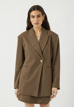 Brun blazer med en struktureret design, enkelt guldtonet hjerteknap og subtile sømdetaljer. Glat stof, let oversized pasform.