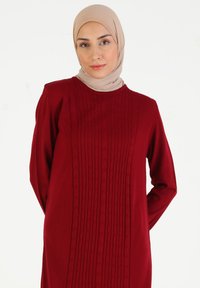 Vestido de punto rojo con mangas largas que presenta patrones verticales texturizados en la parte frontal. Combinado con un hijab beige.
