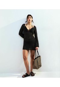 Abito nero a maglia con maniche lunghe e scollatura a V profonda. Una borsa a tracolla a righe e sandali neri completano il look.