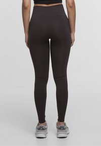 Leggings a vita alta di colore marrone scuro, realizzati in materiale morbido e elastico, con un design aderente e dettagli di cucitura sottili. Indossati con scarpe da atletica grigie.