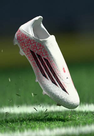 Scarpa da calcio bianca con motivo floreale rosso, strisce nere e tomaia testurizzata, sospesa sopra l'erba verde, con tacchetti visibili sotto.