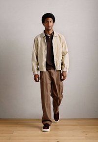 Homme marchant à l'intérieur portant une veste beige, un polo marron, un pantalon marron et des baskets noires sur un sol en bois contre un mur uni.