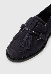 Des mocassins en suédine bleu foncé présentent un design à franges avec un accent noir et un détail de gland, reposant sur une semelle en caoutchouc texturée.