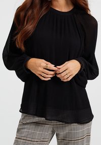 Zwarte geplooide top met lange mouwen en een ronde halslijn, gedragen over gestreepte plaid broek, met subtiele vloei en textuur.