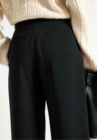 Primo piano di una persona che indossa pantaloni neri su misura e un maglione a treccia color crema, mentre tiene una borsa di pelle nera.