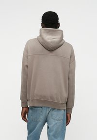 Φούτερ σε ανοιχτό καφέ χρώμα με ευρύχωρη κουκούλα, ριμπ μανσέτες και στρίφωμα. Διαθέτει μινιμ αλιστικό σχέδιο με ραφή στην πλάτη.