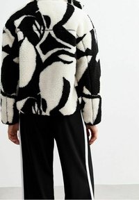 Fleecepusero mustavalkoisella abstraktilla kuviolla, jossa on kauluksellinen pääntie ja suoralinjainen istuvuus. Päällä mustat housut, joissa valkoiset sivuraidat.