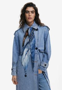 Dunkelblauer Denim-Trenchcoat mit Kragen, langen Ärmeln und zwei Fronttaschen. Mit einem blauen gemusterten Schal, der um den Hals gebunden ist.