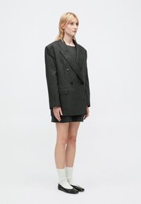Blazer negru, cu două rânduri de nasturi, din țesătură texturată, asociat cu o fustă scurtă. Modelul poartă ciorapi albi și pantofi negri, pe un fundal neutru.