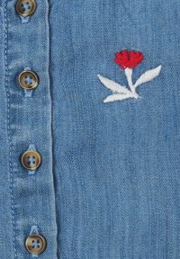 Denim shirt met bruine knopen en een kleine rode geborduurde bloem met groene bladeren op een lichtblauwe stof.