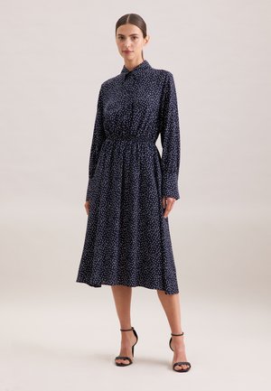 Abito lungo a maniche lunghe blu navy con pois bianchi, colletto a camicia, vita elastica e gonna fluente, abbinato a sandali neri con tacco.