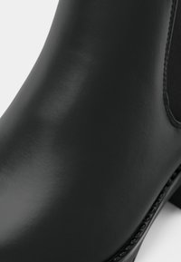 Gros plan sur une botte cheville en cuir noir lisse avec un détail de couture près du panneau élastique latéral et un petit talon.