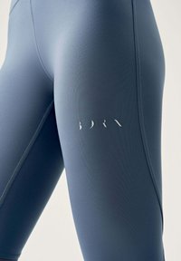 Närbild av blå högmidjade leggings med sömdetaljer och en liten vit logotyp på låret mot en neutral bakgrund.