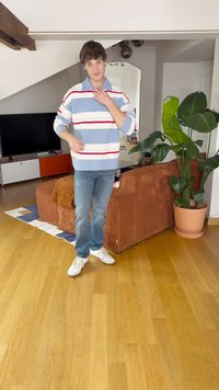 Pruhovaný pletený sveter v modrej, bielej a červenej farbe, kombinovaný s modrými džínami a bielymi teniskami. Interiér s drevenou podlahou a kvetináčom.