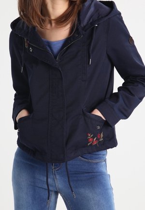 Mujer con chaqueta azul marino con capucha y bordado floral en el bolsillo, camisa azul debajo y vaqueros azules, con las manos en los bolsillos de la chaqueta.