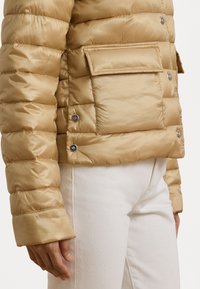 Vaaleanbeige toppatakki kiiltävällä pinnalla, jossa suuri etutasku ja nepparikiinnitys, valkoisten housujen kanssa.