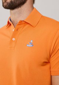 Polo arancione realizzato in tessuto a trama. Presenta un logo ricamato blu sul petto e un colletto a contrasto con bottoni.