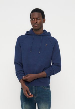 Άνδρας που φοράει μπλε-ναυτικό φούτερ με κουκούλα και μπλε τζιν, στέκεται με τα χέρια ενωμένα μπροστά, με φόντο ένα απλό φωτεινό τοπίο.