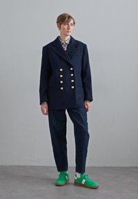 Marineblauer doppelt geknöpfter Mantel mit goldenen Knöpfen, kariertes Hemd, dunkle Hose und grüne Sneakers mit weißen Akzenten. Glatte Stoffqualität, taillierte Passform.