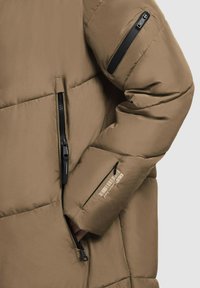 Brūna pildīta jaka ar rāvējslēdzēja kabatām. Gluda tekstūra, melni rāvējslēdzēja akcenti uz piedurknēm, ar zīmola logotipu uz piedurknes netālu no plaukstas.