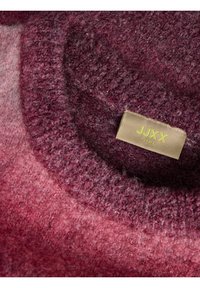 Gros plan sur l'encolure d'un pull en tricot texturé rose et bordeaux avec une étiquette beige portant la mention JJXX GIRL.