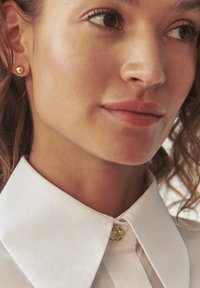 Auksiniai smeigiai auskarai, balta apykaklė, lygus audinys, mygtuko detalė su apvaliu, skaidriu akcentu. Subtili apšvietimas pabrėžia tekstūras.