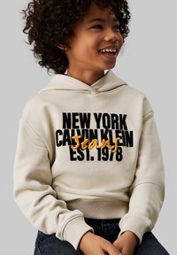 Sudadera crema con texto bordado en negro y amarillo que dice "NEW YORK CALVIN KLEIN Jeans EST. 1978." Presenta un bolsillo tipo canguro y puños acanalados.