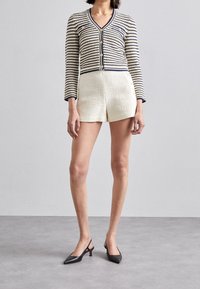 Kobieta w paskowanej, zapinanej kardiganie o kroju cropped, w kremowych, fakturowanych szortach z wysokim stanem oraz czarnych, wskazujących na zapięcie na pięcie szpilkach, stoi na szarym podłożu.