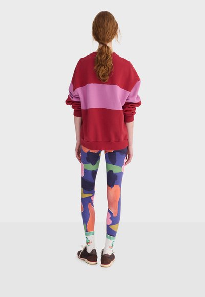 Femme aux longs cheveux roux attachés en queue de cheval, portant un sweat-shirt rayé rouge et rose, des leggings floraux colorés, des chaussettes blanches et des baskets marron, vue de dos.