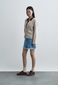 Woman standing wearing beige buttoned cardigan over maroon collared top, blue denim skirt with frayed hem, cream socks, and maroon sneakers.
