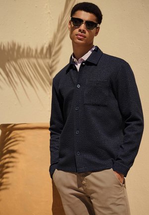Camicia button-up in tessuto blu navy con taschino frontale, indossata sopra una camicia a collare di colore chiaro, abbinata a pantaloni khaki e occhiali da sole.