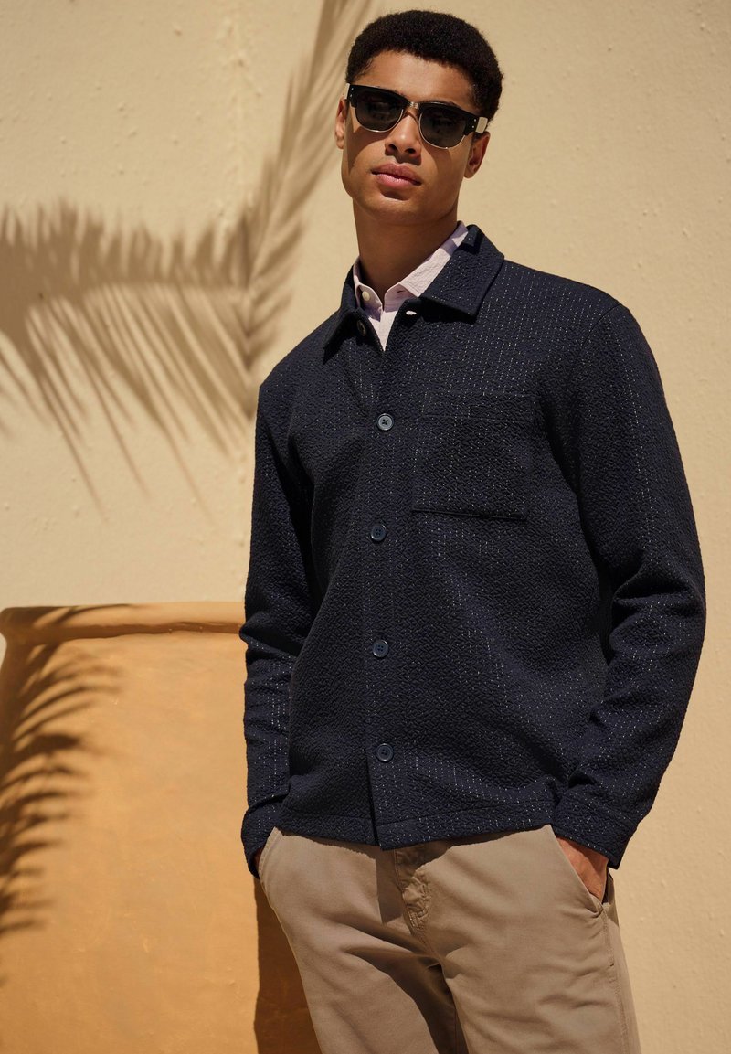 Camicia button-up in tessuto blu navy con taschino frontale, indossata sopra una camicia a collare di colore chiaro, abbinata a pantaloni khaki e occhiali da sole.