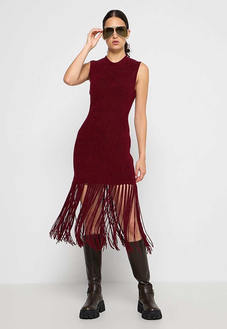 Vestido de punto burdeos con diseño sin mangas y dobladillo con flecos. Lleva botas altas marrones y gafas de sol de gran tamaño.