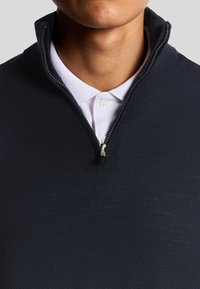 Maglione a collo zip di colore blu navy con tessuto testurizzato, caratterizzato da una cerniera argentata, indossato sopra una camicia bianca con colletto. Vista ravvicinata della zona del collo.