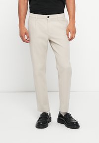 Pantalons en coton beige à coupe droite, avec poches avant et fermeture à un bouton. Associés à des mocassins noirs.