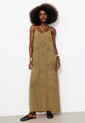 Femme avec une coiffure afro portant une robe maxi olive sans manches avec des motifs en perles, des sandales noires, des bracelets dorés et des colliers superposés.