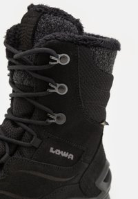Botte de randonnée noire avec tige en suède et textile, col en fausse fourrure, œillets en métal et semelle en caoutchouc épaisse pour une bonne adhérence. Logo de la marque visible.