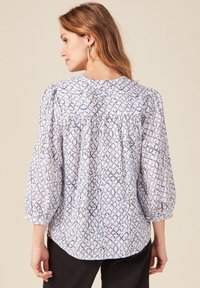 Blusa leggera in bianco con un motivo geometrico blu e nero. Presenta maniche a palloncino e un collo arricciato sul retro.