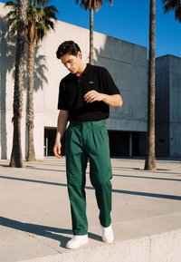 Jeune homme marchant en plein air près des palmiers, portant un polo noir, un pantalon vert et des baskets blanches sous un ciel bleu clair.
