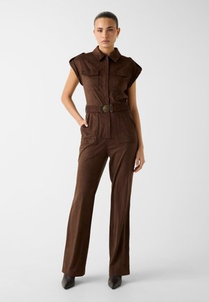 Femme debout, main dans la poche, portant une combinaison marron ceinturée avec manches courtes et chaussures noires pointues sur fond neutre.