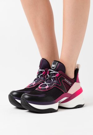 Jambes portant des baskets en mesh Michael Kors noires, violettes et roses avec des semelles blanches épaisses sur un fond blanc.