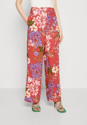 Pantalon large avec fond rouge et grand imprimé floral rose, violet et blanc, porté avec des sandales à talons hauts vertes à lanières.